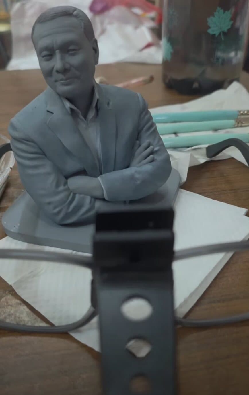 Unpainted grey resin print of a man with folded arms resting on a workbench beside brushes