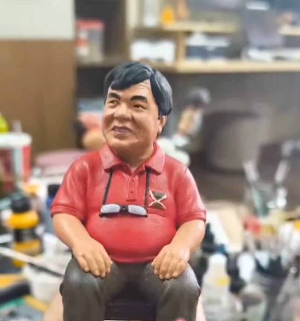 3D printed portrait figurine of a man in a red polo shirt seated in a wooden chair, held above a workbench